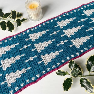 May include: A teal crochet table runner with a white geometric pattern of stylised Christmas trees. The runner has a pink border.
