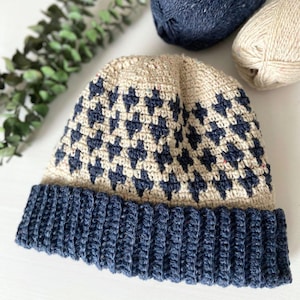 May include: A handmade crochet beanie in a neutral beige color with a geometric navy blue pattern. The brim is a solid navy blue. Two balls of yarn are visible in the background, one navy and one beige.