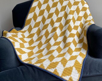May include: A crocheted blanket featuring a repeating geometric diamond pattern in mustard yellow and white. The textile has a textured surface created by the crochet stitches and is finished with a solid dark blue border along the edges.