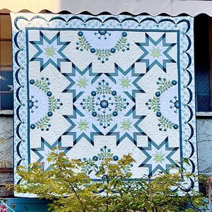 May include: A large, intricately patterned quilt with a cream-colored background. The quilt features a repeating geometric design of blue and green starbursts and floral motifs. The border is a scalloped edge in a darker blue.