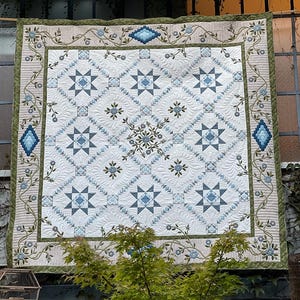 May include: A large, handmade quilt with a detailed star pattern in shades of blue and white. The quilt features a floral border with green and blue accents. The overall design is symmetrical and the quilt is displayed outdoors.