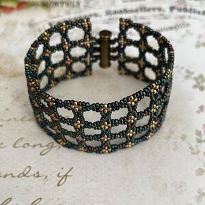 May include: A beaded bracelet with a geometric openwork design. The bracelet features a combination of dark green and gold beads, creating a textured, iridescent appearance. It has a clasp closure and is displayed on a floral patterned surface.