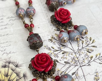 Könnte beinhalten: Ein Set aus Halskette und passenden Ohrringen mit leuchtend roten rosenförmigen Perlen, grauen Perlen mit rosa Blumenmustern sowie filigranen Akzenten aus antikem bronzefarbenem Metall, verbunden durch kleine rote Perlen und Bronzeketten.