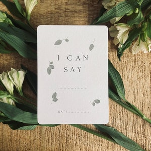 Pode incluir: Um cart&atilde;o branco com o texto "I can say" e uma linha pontilhada para escrever. O cart&atilde;o est&aacute; rodeado de flores brancas e folhas verdes sobre um fundo de madeira.