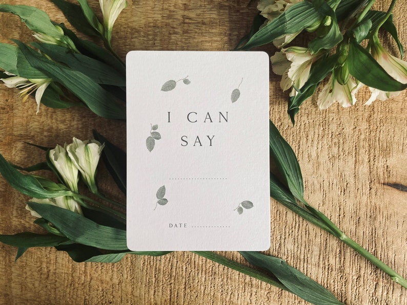 Pode incluir: Um cart&atilde;o branco com o texto "I can say" e uma linha pontilhada para escrever. O cart&atilde;o est&aacute; rodeado de flores brancas e folhas verdes sobre um fundo de madeira.