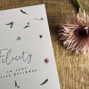 May include: A white card with a floral design and the text "Felicity on your first birthday".