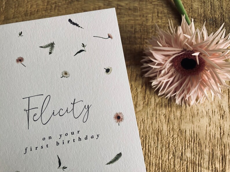 May include: A white card with a floral design and the text "Felicity on your first birthday".