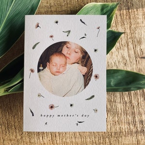 May include: A white Mother's Day card with a circular photo of a mother and baby, surrounded by small floral and leaf illustrations. The words "happy mother's day" are printed at the bottom in a simple font.