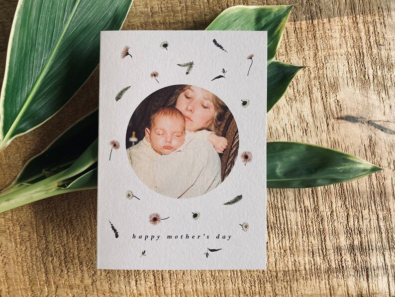 May include: A white Mother's Day card with a circular photo of a mother and baby, surrounded by small floral and leaf illustrations. The words "happy mother's day" are printed at the bottom in a simple font.