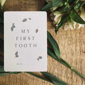 Pode incluir: Um cart&atilde;o branco com o texto "MY FIRST TOOTH" e um espa&ccedil;o para escrever a data. O cart&atilde;o &eacute; decorado com pequenas ilustra&ccedil;&otilde;es de folhas cinzas.