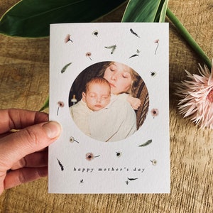 May include: A white Mother's Day card with a circular photo of a mother and baby. The card is decorated with small floral and leaf illustrations and the words "happy mother's day". A pink flower and green leaf are in the background.