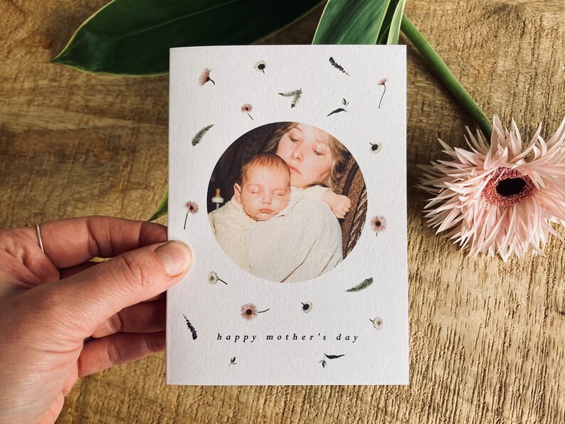May include: A white Mother's Day card with a circular photo of a mother and baby. The card is decorated with small floral and leaf illustrations and the words "happy mother's day". A pink flower and green leaf are in the background.