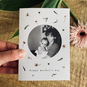 May include: A Mother's Day card featuring a black and white photo of a mother and child within a circular frame. The card is decorated with small floral and leaf illustrations and the words "happy mother's day". A pink gerbera daisy is next to the card.