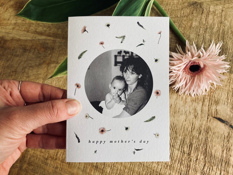 May include: A Mother's Day card featuring a black and white photo of a mother and child within a circular frame. The card is decorated with small floral and leaf illustrations and the words "happy mother's day". A pink gerbera daisy is next to the card.