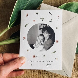 May include: A Mother's Day card featuring a black and white photo of a mother and child within a circle, surrounded by small floral and leaf illustrations. The words "happy mother's day" are printed at the bottom. A beige envelope is included.