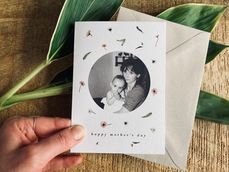 May include: A Mother's Day card featuring a black and white photo of a mother and child within a circle, surrounded by small floral and leaf illustrations. The words "happy mother's day" are printed at the bottom. A beige envelope is included.