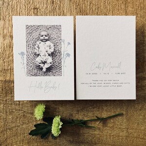Op de afbeelding: Een witte kaart met een zwart-witfoto van een baby die op een kleed ligt. De kaart zegt "Hello Baby!" en heeft een bloemenmotief. Op de achterkant van de kaart staan de naam van de baby, de geboortedatum en het gewicht. Er staat "Hartelijk dank voor al de liefde, wensen, kaarten en cadeaus. Ik ben een heel gelukkig klein baby."
