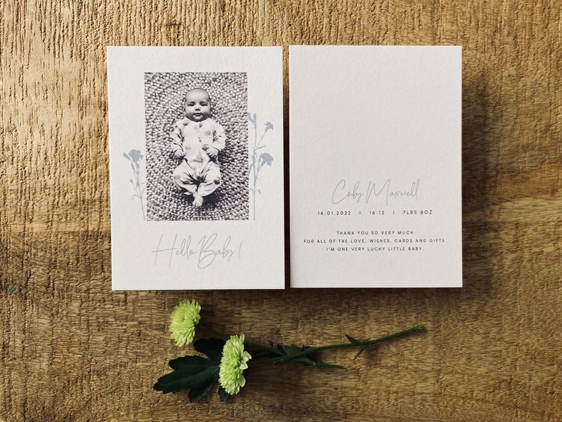 Op de afbeelding: Een witte kaart met een zwart-witfoto van een baby die op een kleed ligt. De kaart zegt "Hello Baby!" en heeft een bloemenmotief. Op de achterkant van de kaart staan de naam van de baby, de geboortedatum en het gewicht. Er staat "Hartelijk dank voor al de liefde, wensen, kaarten en cadeaus. Ik ben een heel gelukkig klein baby."