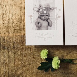 Op de afbeelding: Een witte kaart met een zwart-witfoto van een baby die een hertenhoed draagt. De tekst "Hello Baby!" is in cursief op de kaart geschreven. Een tweede witte kaart met de tekst "When Allen 07.12.2021 02:19" is zichtbaar op de achtergrond. De kaarten liggen op een houten oppervlak.