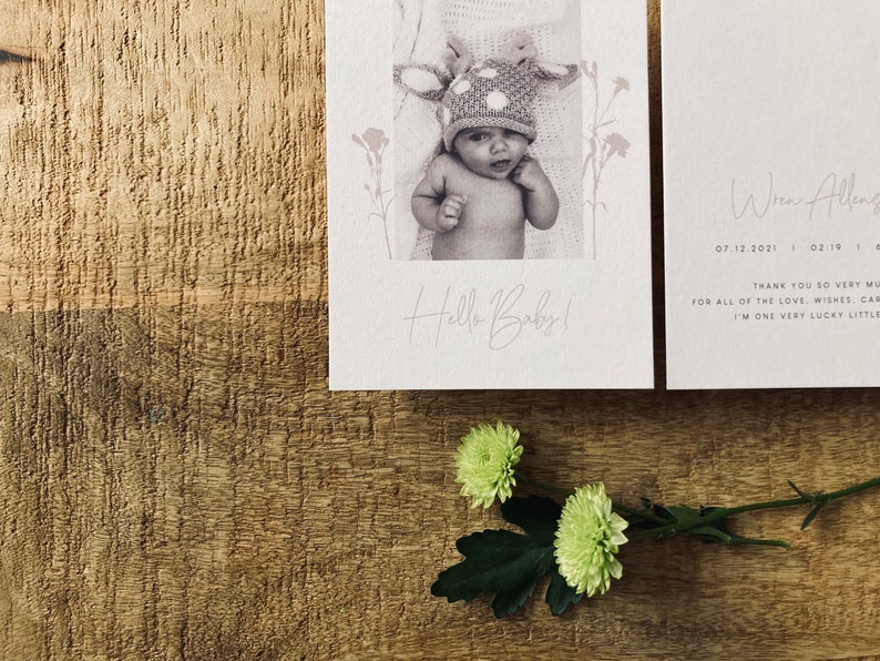 Op de afbeelding: Een witte kaart met een zwart-witfoto van een baby die een hertenhoed draagt. De tekst "Hello Baby!" is in cursief op de kaart geschreven. Een tweede witte kaart met de tekst "When Allen 07.12.2021 02:19" is zichtbaar op de achtergrond. De kaarten liggen op een houten oppervlak.
