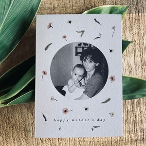 May include: A Mother's Day card with a black and white photo of a mother and child inside a circle, surrounded by small flowers and sprigs. The words "happy mother's day" are printed at the bottom. The card is on a wooden surface with green leaves.