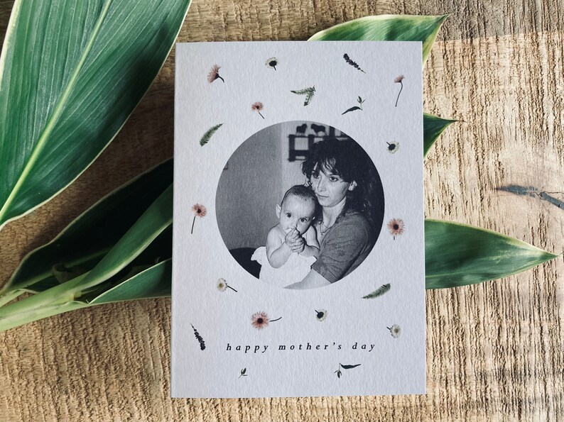 May include: A Mother's Day card with a black and white photo of a mother and child inside a circle, surrounded by small flowers and sprigs. The words "happy mother's day" are printed at the bottom. The card is on a wooden surface with green leaves.