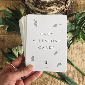 Pode incluir: Um conjunto de cart&otilde;es brancos com o texto "BABY MILESTONE CARDS" em letras pretas. Os cart&otilde;es s&atilde;o decorados com pequenas ilustra&ccedil;&otilde;es de folhas cinzas.