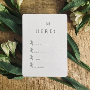 Pode incluir: Um cart&atilde;o branco com o texto "I'M HERE!" e uma lista de informa&ccedil;&otilde;es para preencher: nome, data de nascimento, hora e peso. O cart&atilde;o &eacute; decorado com pequenas ilustra&ccedil;&otilde;es de borboletas cinzas.