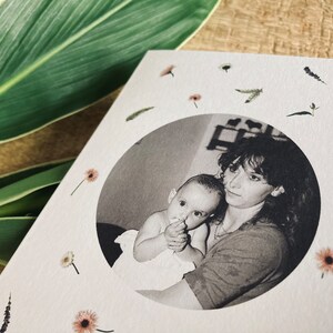 May include: A greeting card featuring a black and white photo of a mother and baby in a circular frame. The card is decorated with small dried flowers and greenery. The background includes green leaves.