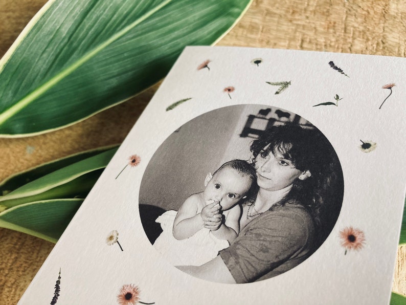 May include: A greeting card featuring a black and white photo of a mother and baby in a circular frame. The card is decorated with small dried flowers and greenery. The background includes green leaves.