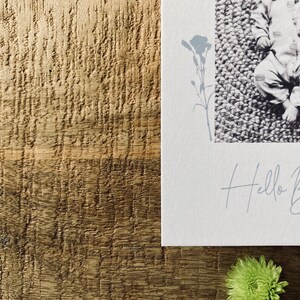 Op de afbeelding: Een witte kaart met een zwart-witfoto van een baby die op een getextureerd oppervlak ligt. De kaart heeft een bloemmotief en de tekst "Hello Baby" in een handschrift.
