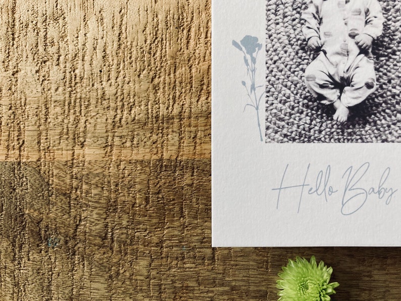 Op de afbeelding: Een witte kaart met een zwart-witfoto van een baby die op een getextureerd oppervlak ligt. De kaart heeft een bloemmotief en de tekst "Hello Baby" in een handschrift.