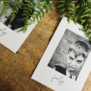 May include: Two white cards with black and white photos and the word "forty" written in cursive on each card. The cards are on a wooden surface with green fern leaves.