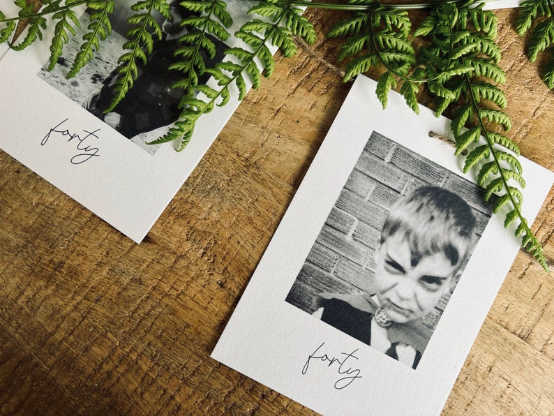 May include: Two white cards with black and white photos and the word "forty" written in cursive on each card. The cards are on a wooden surface with green fern leaves.