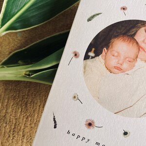 May include: A greeting card with a circular photo of a mother and baby, surrounded by floral and feather illustrations. The words "happy mom" are printed at the bottom. Green and white leaves are visible in the background.