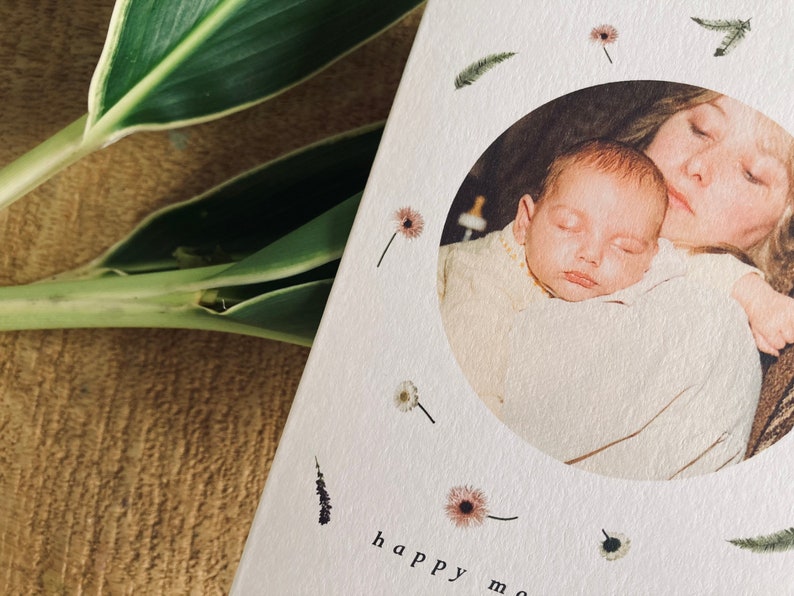 May include: A greeting card with a circular photo of a mother and baby, surrounded by floral and feather illustrations. The words "happy mom" are printed at the bottom. Green and white leaves are visible in the background.