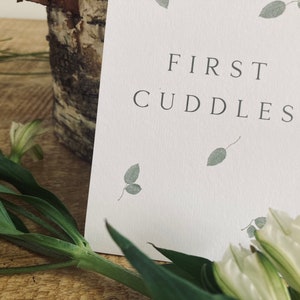 Pode incluir: Um cart&atilde;o branco com o texto "FIRST CUDDLES" em letras pretas. O cart&atilde;o &eacute; decorado com pequenas folhas verdes.