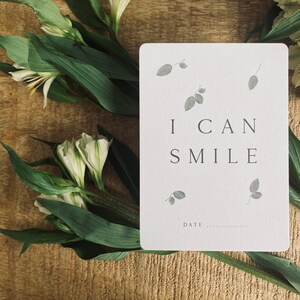 Pode incluir: Um cart&atilde;o branco com o texto "I CAN SMILE" e um pequeno desenho de folhas cinzas. O cart&atilde;o est&aacute; sobre uma superf&iacute;cie de madeira com flores brancas e folhas verdes.