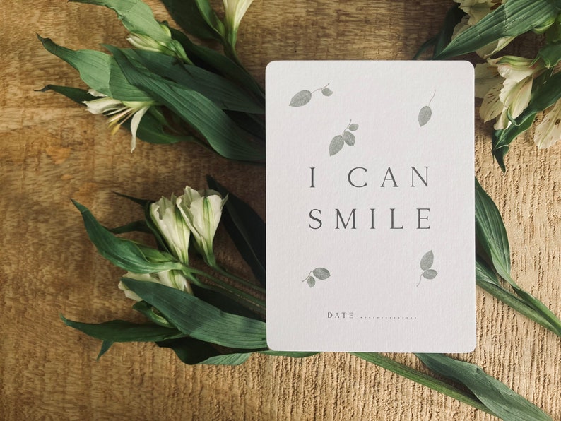 Pode incluir: Um cart&atilde;o branco com o texto "I CAN SMILE" e um pequeno desenho de folhas cinzas. O cart&atilde;o est&aacute; sobre uma superf&iacute;cie de madeira com flores brancas e folhas verdes.