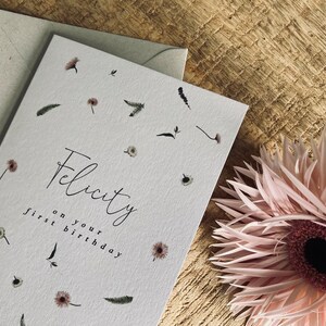 May include: A white card with a floral design and the text "Felicity on your first birthday".