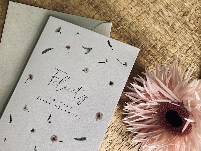 May include: A white card with a floral design and the text "Felicity on your first birthday".