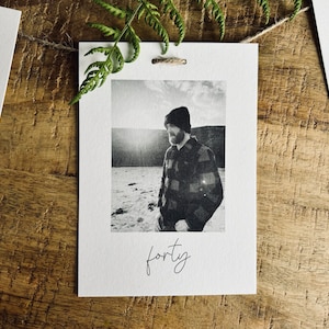 May include: A black and white photo of a person wearing a plaid shirt and a beanie hat. The photo is printed on a white card with the word "forty" written in cursive below the photo.