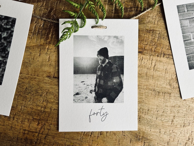 May include: A black and white photo of a person wearing a plaid shirt and a beanie hat. The photo is printed on a white card with the word "forty" written in cursive below the photo.