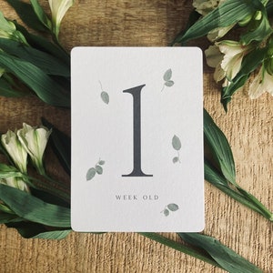 Pode incluir: Um cart&atilde;o branco com um n&uacute;mero um preto e o texto "week old" impresso nele. O cart&atilde;o &eacute; cercado por folhas verdes e flores brancas.