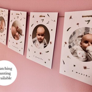 May include: A set of white photo cards with a circular cutout for a photo of a baby. Each card has a floral design and the text "x months" printed on it. The cards are strung together with twine.