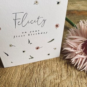 May include: A white card with the text "Felicity on your first birthday" in black script. The card is decorated with small, white flowers and green leaves.