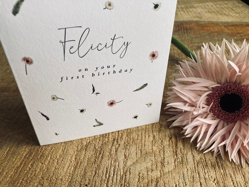 May include: A white card with the text "Felicity on your first birthday" in black script. The card is decorated with small, white flowers and green leaves.