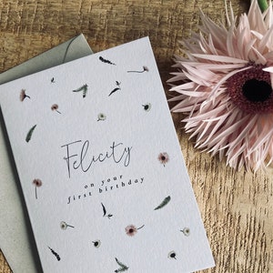 May include: A white card with a floral pattern and the text "Felicity on your first birthday".