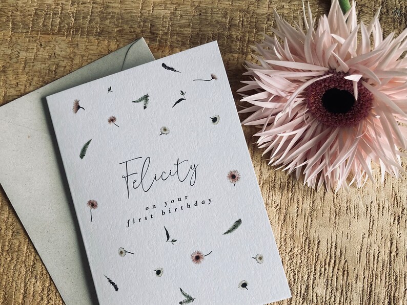 May include: A white card with a floral pattern and the text "Felicity on your first birthday".