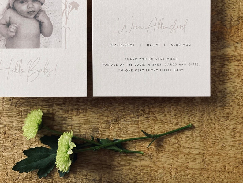 Op de afbeelding: Een witte kaart met de tekst "Hello Baby!" in cursief schrift. De kaart ligt naast een witte kaart met de tekst "Wren Allengford" in cursief schrift. De tekst "07.12.2021 | 02:19 | 6LBS 9OZ" is onder de naam gedrukt. De tekst "Bedankt voor al de liefde, wensen, kaarten en cadeaus. Ik ben een heel gelukkig klein baby" is onder de datum en tijd gedrukt.